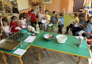 dzieci siedzą wokół stolika i podnoszą rękę w górę