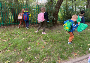 Na zdjęciu kilkoro dzieci z plecakami przeszukuje teren przy ogrodzeniu, zbierając śmieci do zielonych worków.