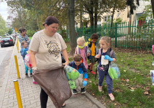 Na zdjęciu widać, jak dzieci i opiekunka aktywnie zbierają śmieci z trawnika przy ulicy. Dzieci podnoszą różne drobne odpady i wkładają je do worków.