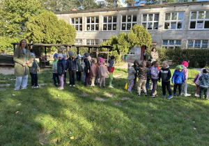 grupa dzieci i kilku dorosłych stoją na trawniku w ogrodzie przedszkolnym