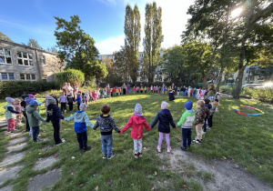 grupa dzieci i kilku dorosłych stoją na trawniku w ogrodzie przedszkolnym i trzymają się za ręce