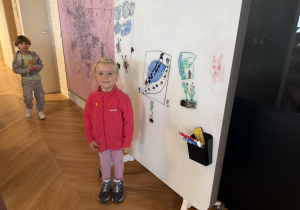 dziewczynka stoi obok tablicy z rysunkami a z tyłu stoi chłopiec