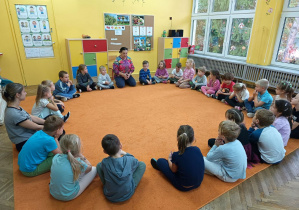Grupa dzieci siedzi w kole na dywanie z panią prowadzącą