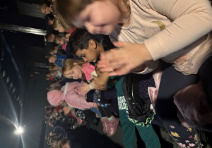 Z boku sali widać grupę dzieci siedzących w fotelach kinowych. Dzieci są uśmiechnięte, klaszczą i reagują z entuzjazmem na to, co dzieje się na scenie. Mają na sobie kolorowe ubrania, kurtki i bluzy. W tle widać kolejne rzędy siedzeń i ciemne ściany sali teatralnej. Atmosfera jest radosna i pełna energii.