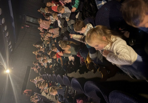 Widzowie siedzą w rzędach w sali teatralnej. W większości to dzieci, które z zainteresowaniem patrzą w stronę sceny. Niektóre klaszczą, inne śmieją się lub rozmawiają z kolegami. W tle widać dorosłych opiekunów. Oświetlenie jest przygaszone, a punktowe światło pada na publiczność.