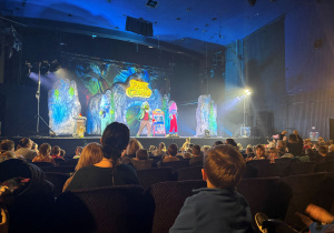 W dużej sali teatralnej trwa przedstawienie zatytułowane „Park Czarów”. Na scenie, ozdobionej kolorowymi dekoracjami przypominającymi skalne bramy i jaskinie, stoją aktorzy w barwnych kostiumach. Światła sceniczne tworzą magiczną atmosferę. Przed sceną siedzi wielu widzów – głównie dzieci – oglądających spektakl z zaciekawieniem.