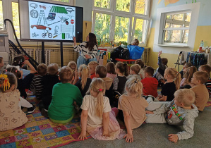 grupa dzieci siedzi na dywanie i patrzy w stronę ekranu i pani prowadzącej