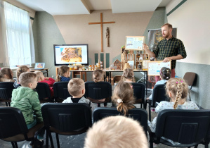 Dzieci siedzą na krzesłach w sali i słuchają prowadzącego, który stoi przed nimi