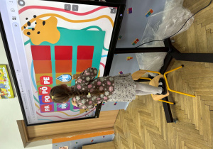 Dziewczynka w kwiecistej sukience stoi na krześle, aby dosięgnąć do dużego ekranu dotykowego. Na ekranie widoczna jest gra z kolorowymi sylabami – „PA”, „PO”, „PÓ”, „PE”. Dziecko skupione dotyka wybranego pola. W sali widać drewnianą podłogę, jasne ściany i kolorowe dekoracje.