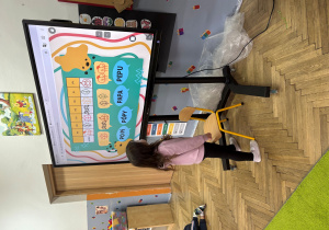 Dziewczynka w różowej bluzie stoi przed dużym ekranem dotykowym, na którym wyświetlona jest kolorowa gra językowa z sylabami i symbolami. Dziecko dotyka ekranu, ucząc się poprzez zabawę. Obok stoi małe krzesełko, a w tle widać fragmenty przedszkolnej sali.