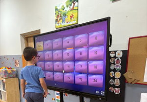 Chłopiec stoi na krześle przed dużym ekranem dotykowym w przedszkolu. Na ekranie widoczna jest kolorowa gra edukacyjna z ponumerowanymi polami. Chłopiec dotyka jednego z kwadratów, biorąc aktywny udział w zabawie. Wokół widać elementy sali – drewnianą podłogę, kolorowe dekoracje i dziecięce meble.