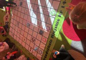 Dzieci siedzą wokół tej samej maty z kratkami. Na macie znajdują się ilustracje zwierząt i innych rzeczy, a na brzegu widnieje napis „Przedszkole Miejskie Nr 144”. W centrum maty są rozłożone obrazki m.in. z rybą, ptakiem i kotem. Dzieci trzymają w dłoniach kolorowe piłeczki lub kubeczki, czekając na swoją kolej w zabawie.