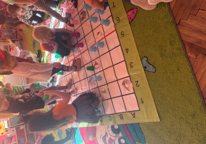 Grupa dzieci bawi się na tej samej macie z kratkami. Dzieci stoją i klęczą, trzymając kolorowe plastikowe kubeczki, które ustawiają w różnych miejscach na macie. Na kratkach nadal widoczne są obrazki zwierząt i przedmiotów. Całość odbywa się w kolorowej sali przedszkolnej, na dywanie z wzorami i postaciami.