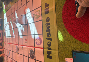 robot stoi na macie w kratkę z obrazkami, a wokół siedzą dzieci. Na pierwszym planie widać kartonik z narysowanymi strzałkami i tablet, który prawdopodobnie służy do sterowania robotem. Na macie widoczne są obrazki z napisem „bird” i „cat”, czyli ptak i kot. Światło słoneczne wpada przez okno, tworząc jasne plamy na macie.