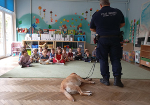 Strażnik miejski z psem na smyczy i siedząca grupa dzieci