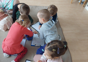 dzieci siedzą na dywanie i kolorują kredkami kolorowankę