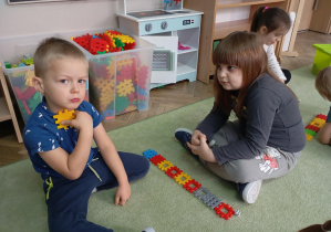 dzieci siedzą na dywanie, przed nimi leżą ułożone z kolorowych klocków rzędy