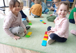 dzieci siedzą na dywanie, przed nimi leżą ułożone z kolorowych klocków rzędy