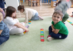 dzieci siedzą na dywanie, przed nimi leżą ułożone z kolorowych klocków rzędy