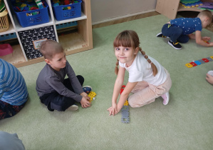 dzieci siedzą na dywanie, przed nimi leżą ułożone z kolorowych klocków rzędy