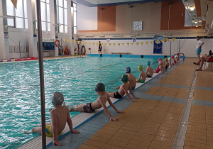 grupa dzieci siedzi na brzegu basenu