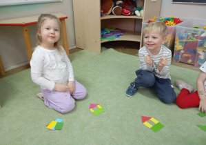 dziewczynka i chłopiec siedzą na dywanie przed nim leżą figury geometryczne
