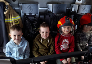 dzieci w wozie strażackim gdzie dwoje ma na sobie hełmy strażackie