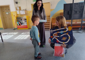 pani dyrektor kuca w stronę dziecka z grą planszową a druga pani trzyma nagrodę