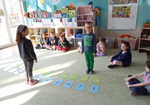 chłopiec w zielonych spodniach i bluzce, a naprzeciwko niego dziewczynka. Na podłodze między nimi leżą kartki z cyframi oraz karty z kropkami – wygląda to na zabawę edukacyjną związaną z nauką liczenia