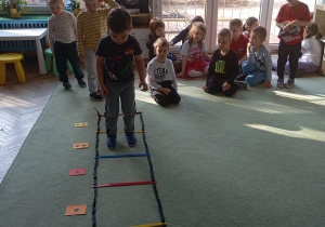 W centrum zdjęcia znajduje się chłopiec, który przechodzi po drabince koordynacyjnej rozłożonej na podłodze. Drabinka ma kolorowe szczebelki, a obok niej leżą kartki z kropkami – prawdopodobnie służące do liczenia lub jako element zadania ruchowo-edukacyjnego