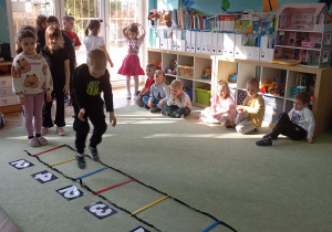 W centrum zdjęcia znajduje się chłopiec, który przechodzi po drabince koordynacyjnej rozłożonej na podłodze. Drabinka ma kolorowe szczebelki, a obok niej leżą kartki z kropkami – prawdopodobnie służące do liczenia lub jako element zadania ruchowo-edukacyjnego