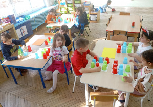 dzieci siedzą przy stolikach.Każde z nich ma przed sobą kolorowe kubeczki