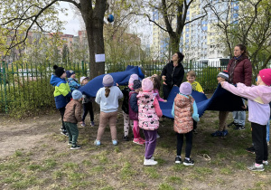 dzieci stoją w ogrodzie przedszkolnym, trzymają chustę w górze