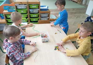 dzieci siedzą przy stoliku i nawlekają koraliki na sznurek