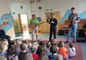 trzech muzyków na scenie i dzieci siedzące twarzami do muzyków