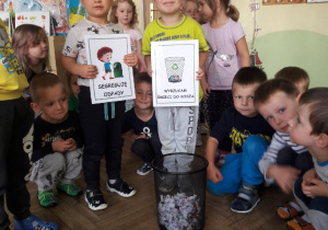 grupa dzieci otacza kosz ze śmieciami a dwoje dzieci stoi i trzyma obrazki z napisami "Wyrzucam śmieci" oraz "Segreguję odpady