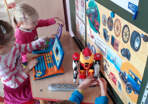 dwie dziewczynki bawią się laptopami zabawkowymi, widoczny robot, pilot i obrazki z urządzeniami