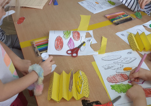 obrazki i karty pracy z produktami, widoczne ręce dziecka które koloruje oraz po lewo widoczny profil dziewczynki która smaruje klejem papier