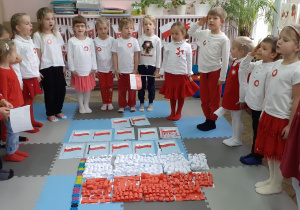 grupa dzieci stoi w półkolu otaczając dużą flagę białoczerwona wykonaną z papieru i śpiewa hymn