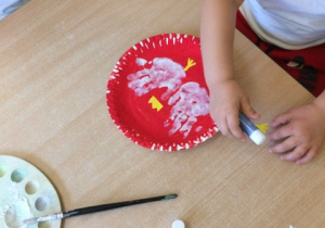 widoczne rączki dziecka posługującego się klejem oraz dwa papierowe godła z talerzyków