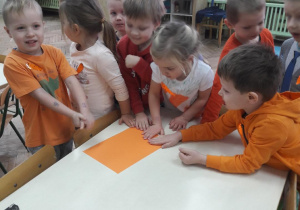 grupka dzieci stojąc przy stole dotyka pomarańczowej kartki