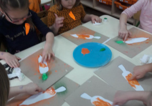 dzieci malują paluszkami umoczonymi w farbie sylwetę marchewki przy stole