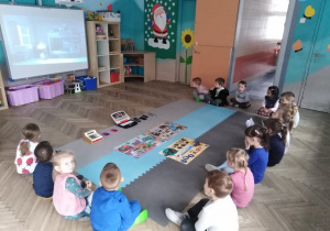 dzieci siedzą wokół maty przed tablicą interaktywną na której sa różne przedmioty i ilustracje