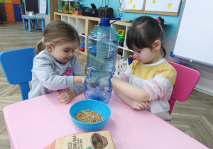 dwie dziewczynki siedzą przy stoliku i rzucają ziarenka do plastikowej butelki