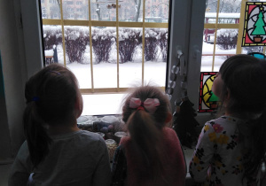 trójka dzieci stojąca przodem do okna patrzy na karmnik który wisi za oknem