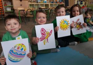 czworo dzieci siedzi na macie i pokazuje obrazki wielkanocne i wiosenne