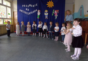 grupa dzieci z nauczycielką stoi w półkolu i pokazuje rękoma gesty na tle dekoracji na pasowanie