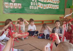 Grupa dzieci siedzi na dywanie. Każde dziecko trzyma w ręku wstążki w kolorze białym i czerwonym.