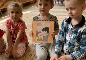 Troje dzieci siedzi na dywanie . Chłopiec trzyma przed tekst z ilustracją