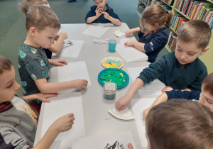 Grypa dziec siedzy przy stoliku i naklejają na papier kawałki kolorowego papieru
